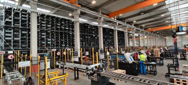 Duża hala produkcyjna stali, z wysokimi regałami pełnymi długich elementów stalowych, systemem przenośników i maszyn do obróbki; w środku pracownik w kamizelce odblaskowej obsługuje urządzenie, a nad całością widać suwnice i konstrukcję przemysłową.