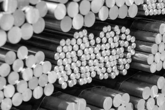 Stos metalowych prętów o okrągłym przekroju, ułożonych poziomo w wiązki. Ich końcówki tworzą gęsty, powtarzalny wzór jasnych kół o różnych średnicach, a powierzchnia prętów jest gładka i lekko połyskująca.