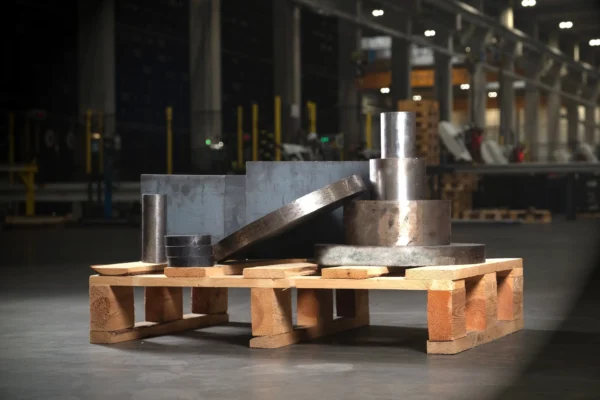 Stalowe walce i cylindry ułożone na drewnianej palecie w hali magazynowej.